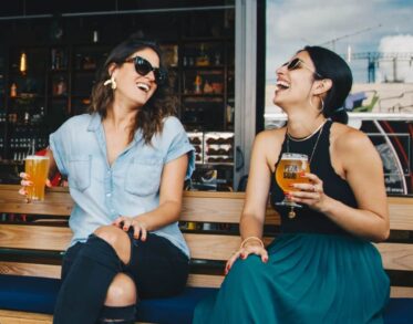 Two women laughing and enjoying drinks on a wooden bench at a bar, with one holding a glass of beer