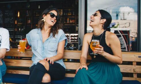 Two women laughing and enjoying drinks on a wooden bench at a bar, with one holding a glass of beer