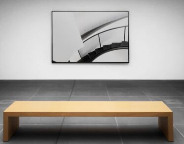 Minimalist art gallery with a black and white photo of a staircase on the wall and a wooden bench in front