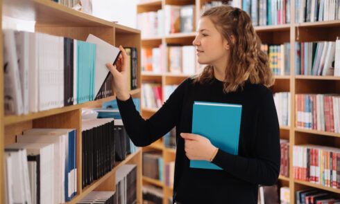 Teenager girl picking a book on a library shelf